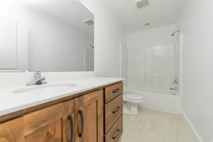 Wood-finish vanity with matte black hardware, white countertop with integrated sink, chrome faucet, white tub and shower combination with chrome fixtures, and light-toned tile flooring