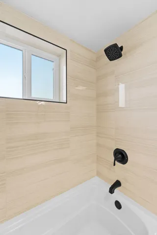 Bathtub with light-toned tiled surround, matte black fixtures, and an integrated window