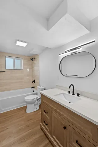 Wood-finish flooring, light wood vanity with a white countertop, modern black-framed oval mirror, and a tub with tiled surround