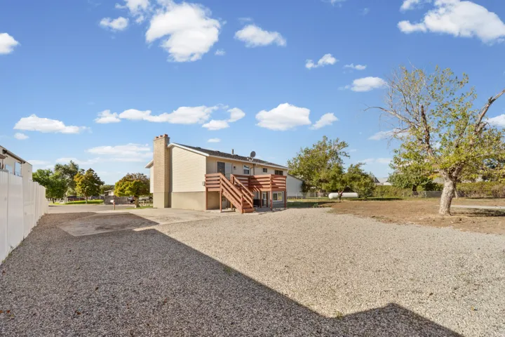 Gravel surface area, rear deck with wood railing, tan exterior siding, brick chimney, and a white privacy fence