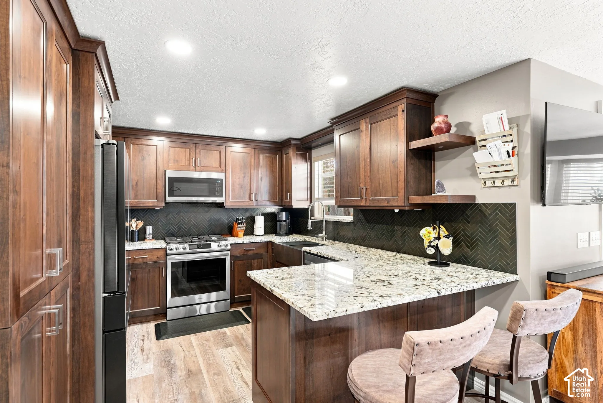 Kitchen featuring cherrywood cabinetry, stainless steel appliances, speckled stone-finish countertops, herringbone-patterned tile backsplash, and recessed lighting