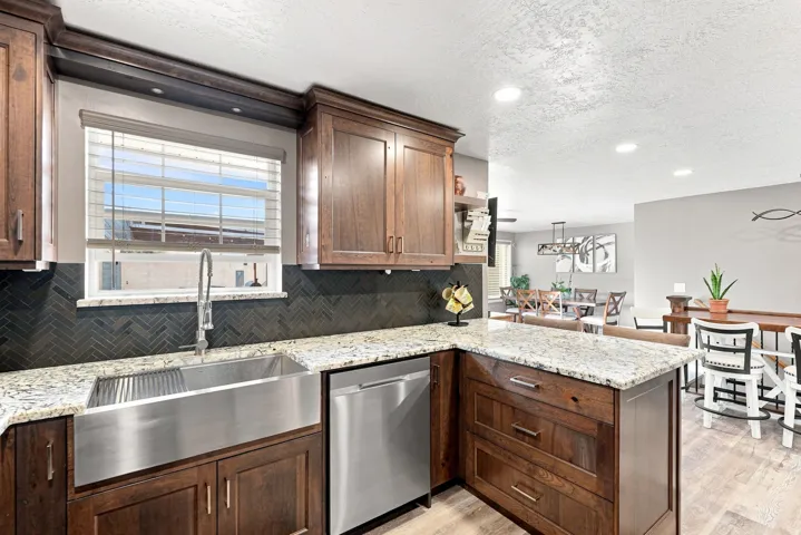 Kitchen featuring wood cabinetry, light-toned countertops, a stainless steel apron front sink, a dark herringbone tile backsplash, and recessed lighting