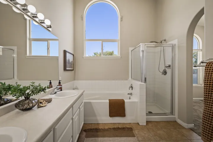 Arched window with ample natural light, spacious built-in tub, separate glass-enclosed shower, dual-sink vanity with under-cabinet storage, and neutral tile flooring