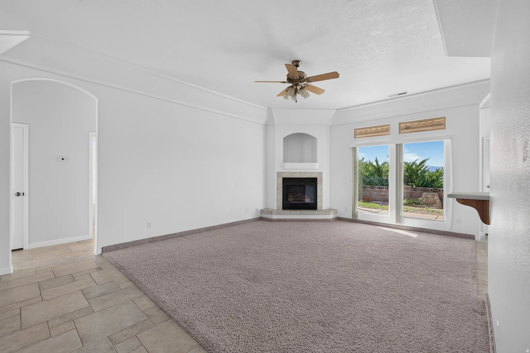 Corner fireplace with a tile surround, large windows, ceiling fan, and arched entryway