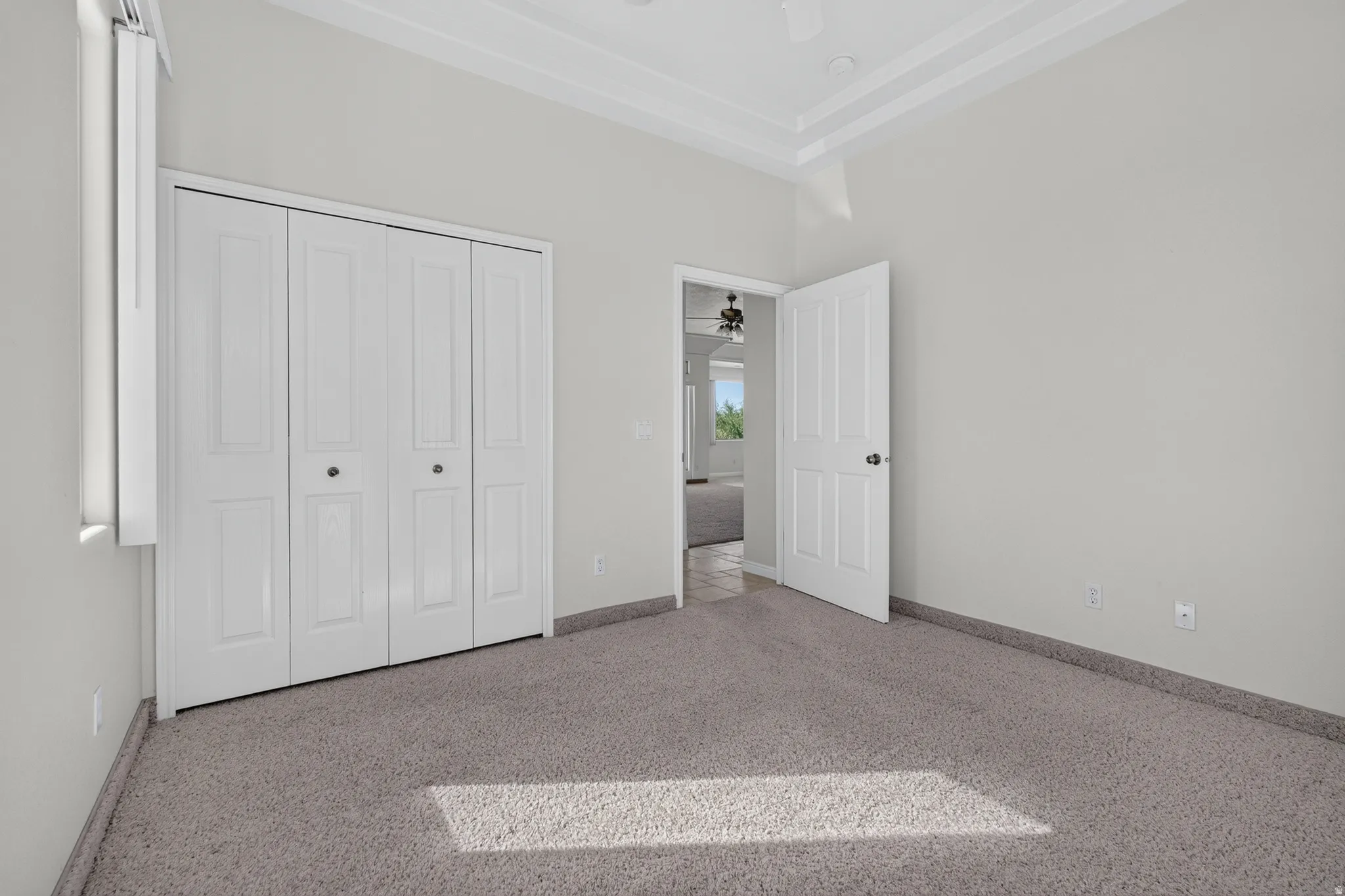 Tray ceiling with crown molding, ceiling fan, bi-fold closet doors, wall-to-wall carpeting, and neutral wall paint
