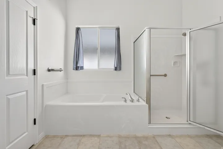 Soaking tub with deck-mounted fixtures, separate enclosed shower with grab bar, frosted window, and tile flooring