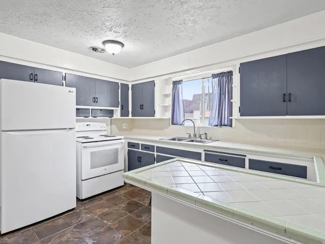 Kitchen featuring dark cabinetry, white appliances, a double basin sink, tile countertops, and stone-finish flooring