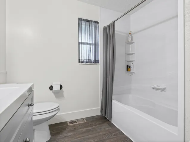 White vanity with integrated sink, toilet, and a combined bathtub and shower with built-in shelving, all complemented by wood-finish flooring