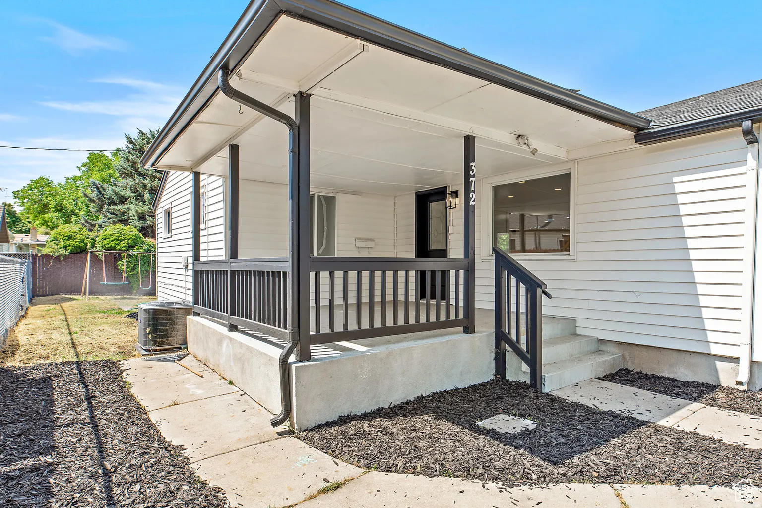 Front exterior featuring white horizontal siding, a covered porch with dark railings and support posts, an entry door with sidelights, and concrete steps