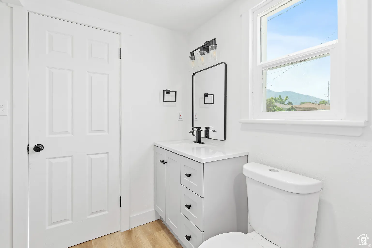 Bathroom featuring a single window, a light wood-finish floor, a vanity with a white countertop and black fixtures, a rectangular framed mirror, and a white toilet