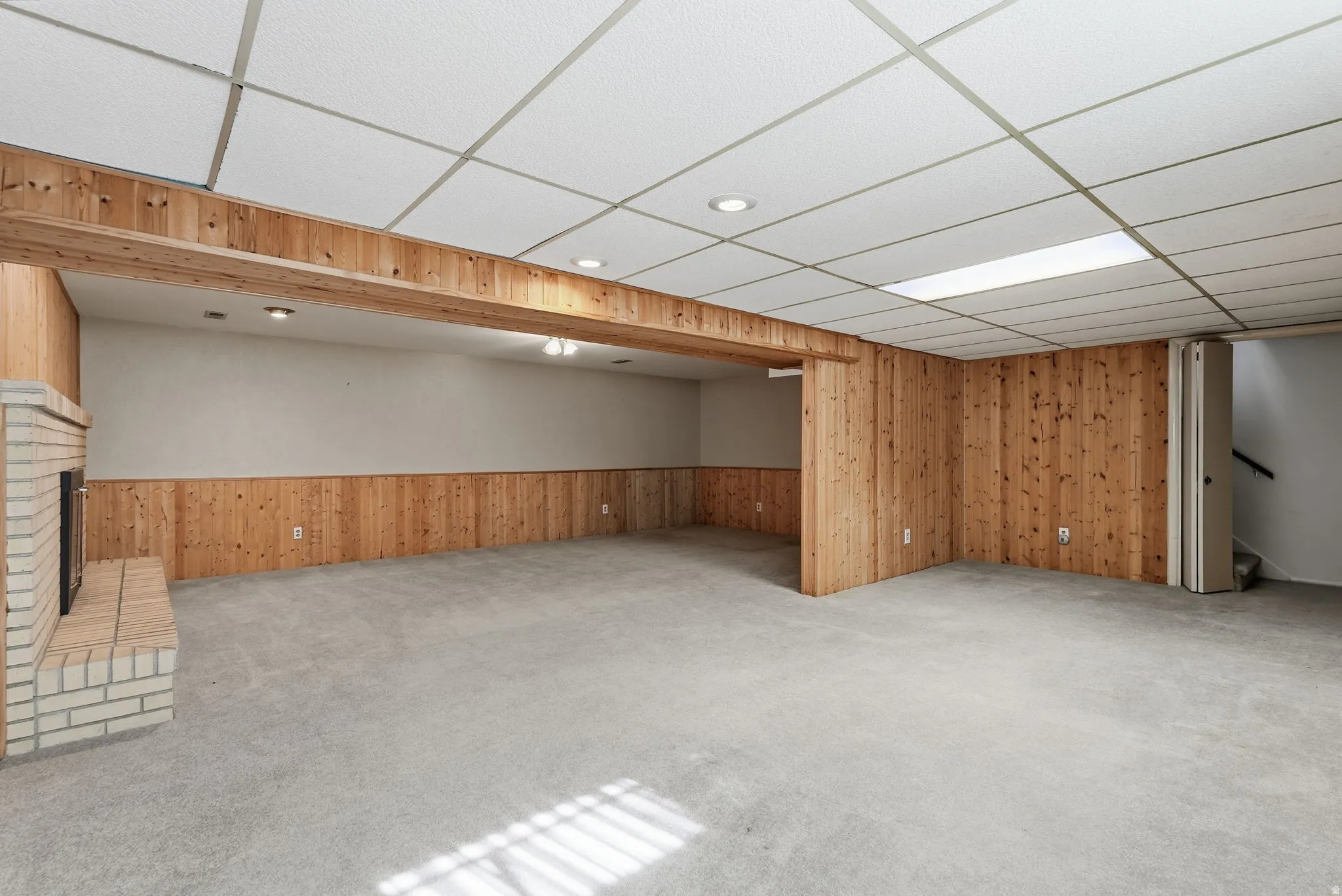 Expansive carpeted area featuring a brick fireplace, wood paneling, and a drop ceiling with recessed lighting