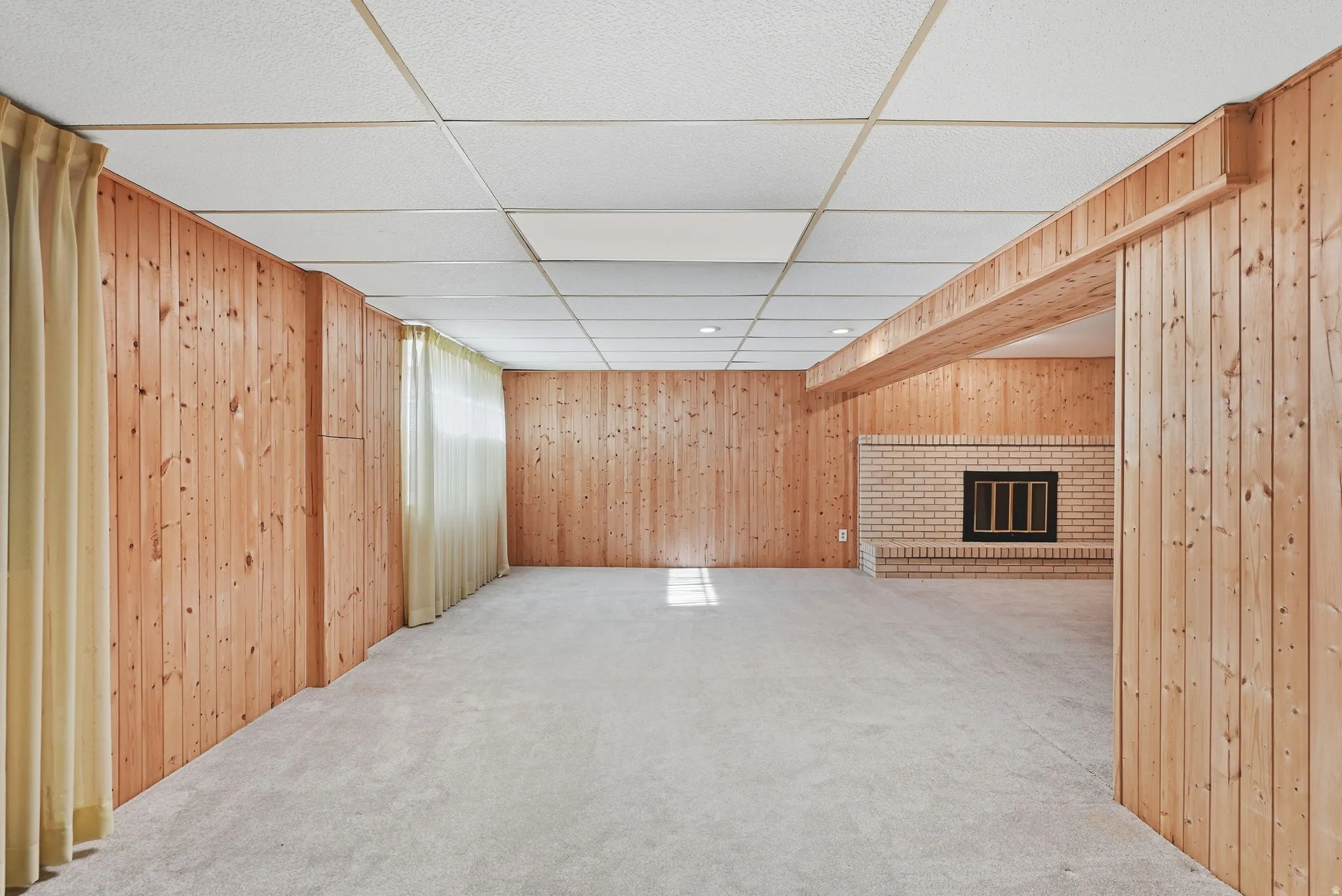 Recessed lighting, drop ceiling, wood-paneled walls, brick fireplace with black grate, and light-toned carpeting