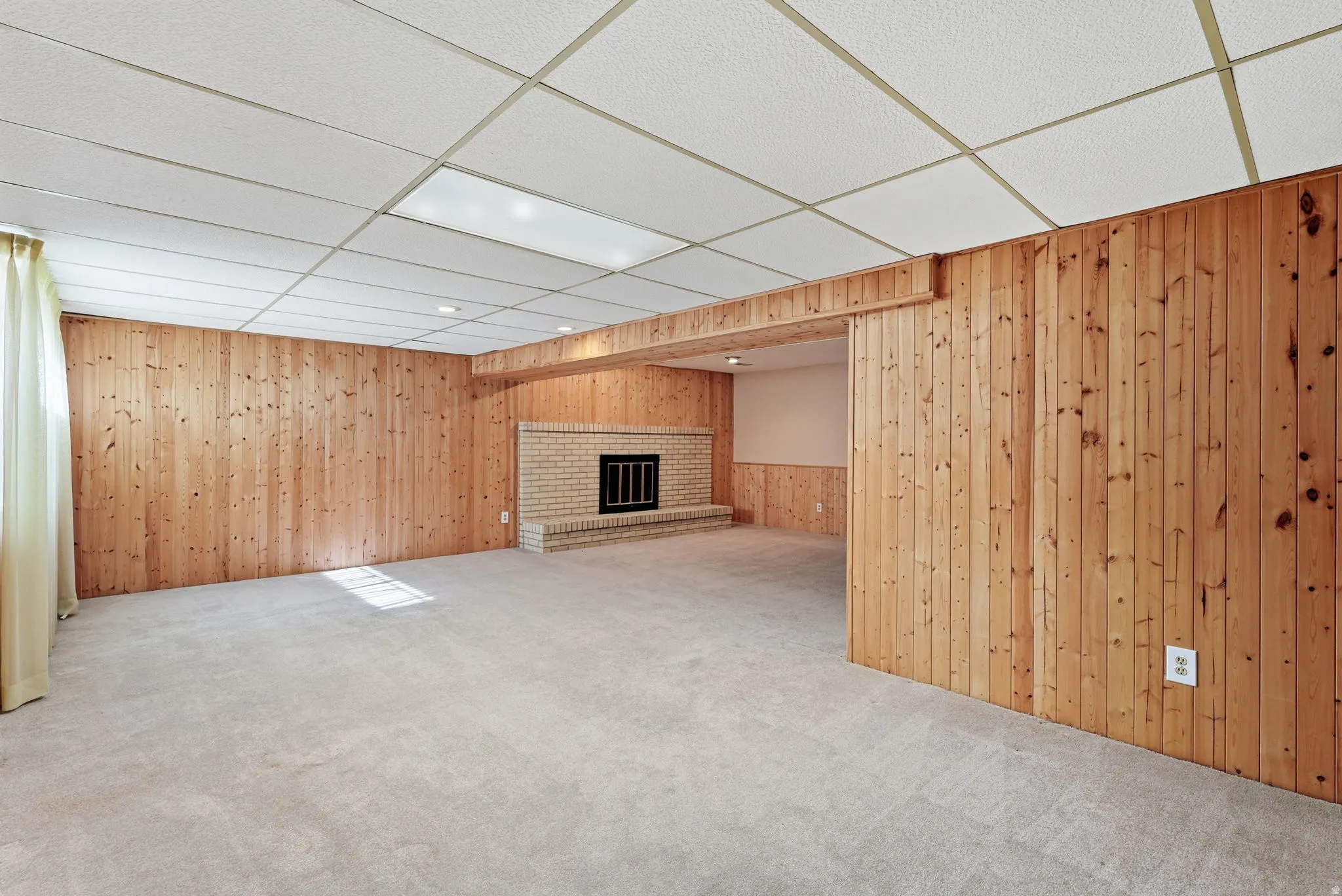 Carpeted flooring, wood-paneled walls, brick fireplace with black grate, drop ceiling with recessed lighting, and sheer curtains