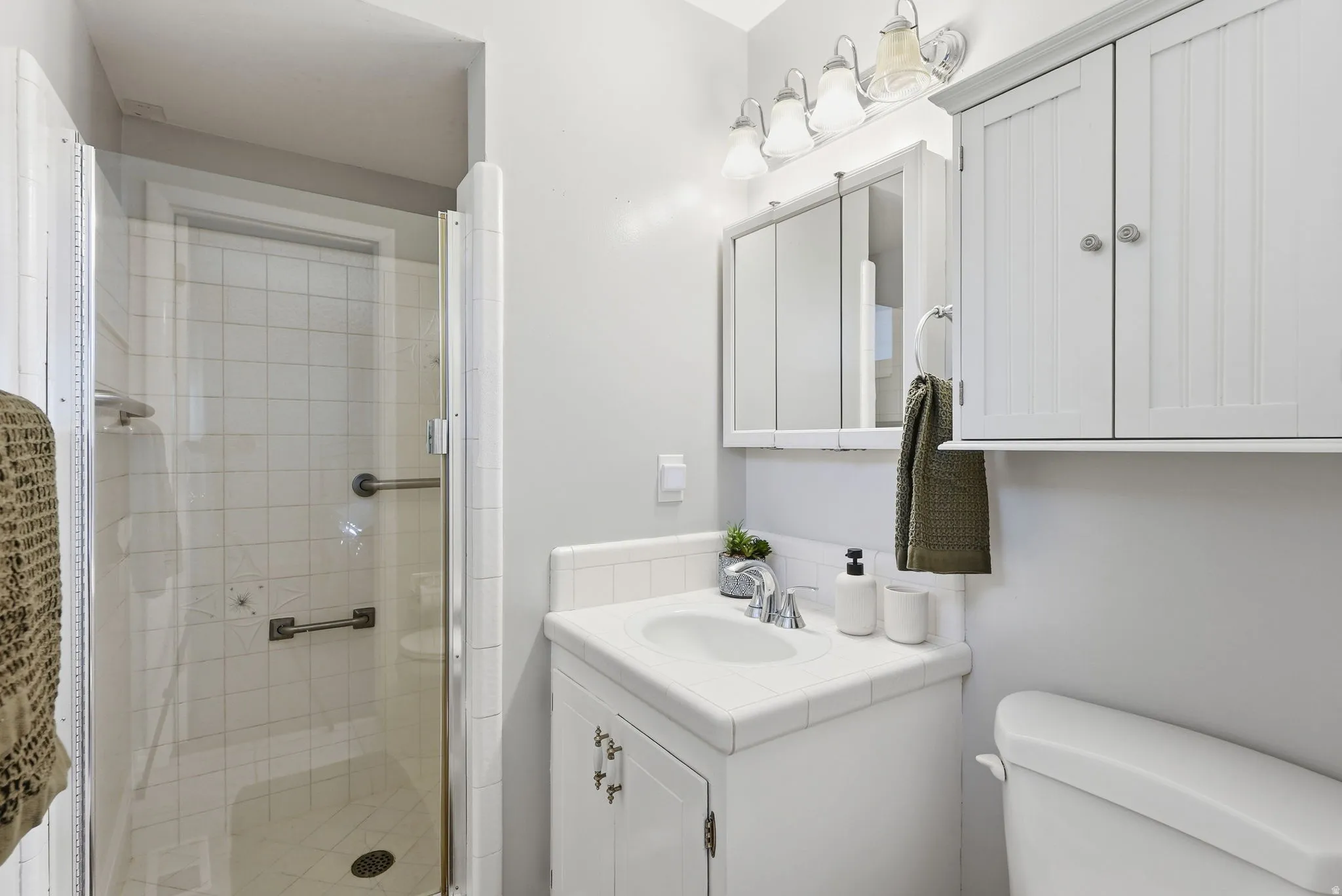 Glass-enclosed shower with white tiled walls, white vanity with integrated sink, chrome faucet, wall-mounted medicine cabinet, and overhead built-in storage