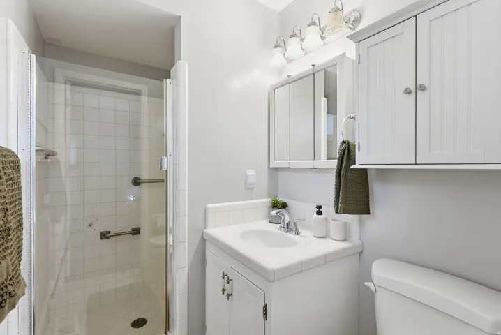 Glass-enclosed shower with white tiled walls, white vanity with integrated sink, chrome faucet, wall-mounted medicine cabinet, and overhead built-in storage