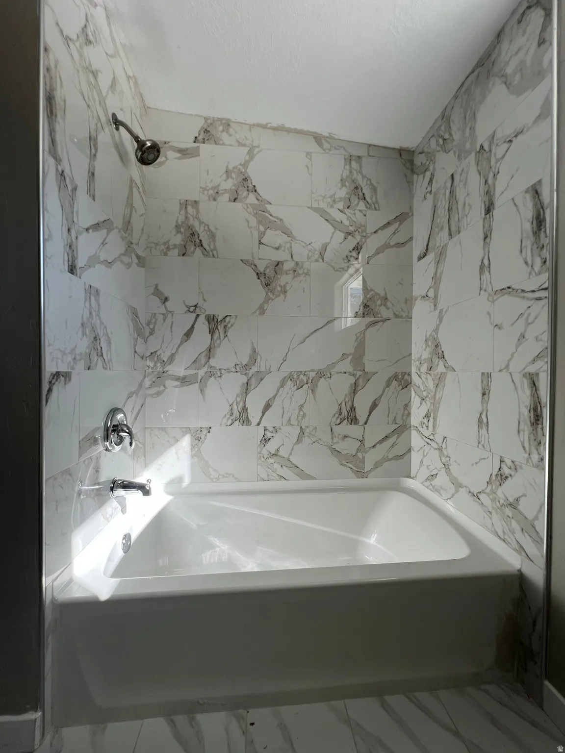 Bathtub with chrome fixtures, tiled surround featuring white and grey marbling, and a built-in shower niche