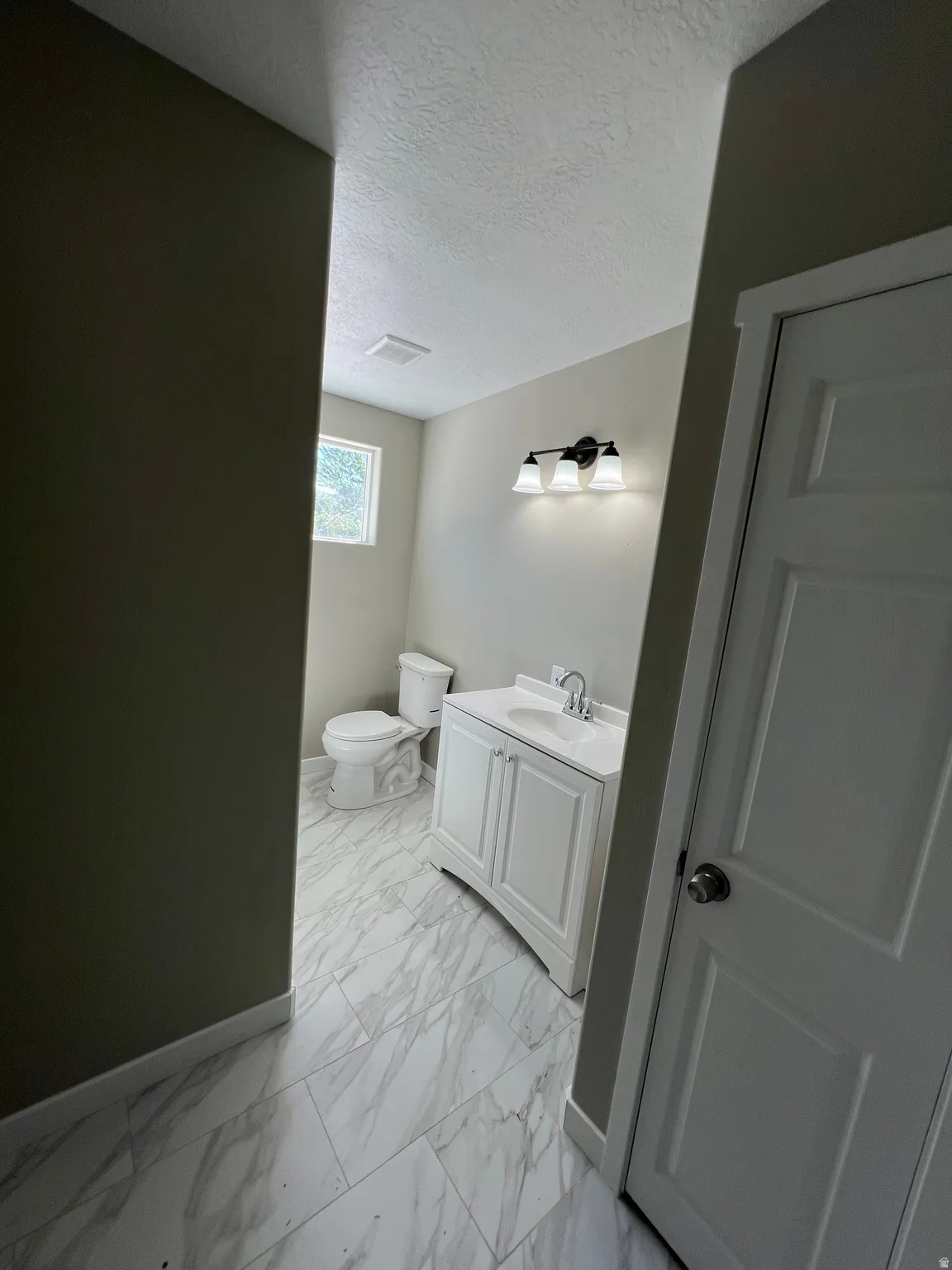 White vanity with integrated sink, chrome faucet, wall-mounted light fixture, white toilet, and marble-look tile flooring