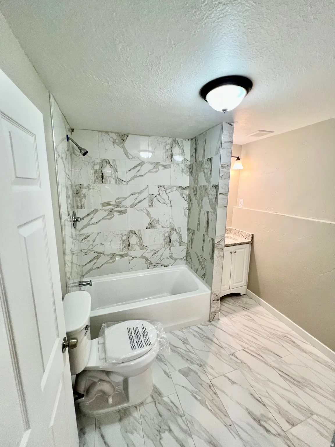 White marble-patterned tile flooring, white vanity with stone-finish countertop, white bathtub with a tile surround, and a white toilet
