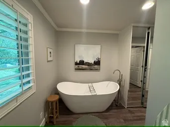Freestanding soaking tub with floor-mounted faucet, plantation shutters, and wood-finish flooring