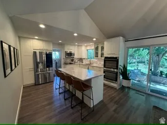 Vaulted ceiling with recessed lighting, wood-finish flooring, white cabinetry with built-in stainless steel double ovens and refrigerator, kitchen island with stone countertop, and sliding glass patio doors