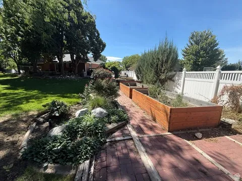 Expansive backyard featuring a manicured lawn, brick paver pathway, multiple raised garden beds, established landscaping, and a white privacy fence