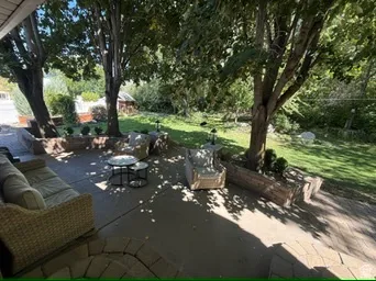Expansive patio with paver and concrete flooring, mature trees providing natural shade, stone retaining walls, and a manicured lawn area