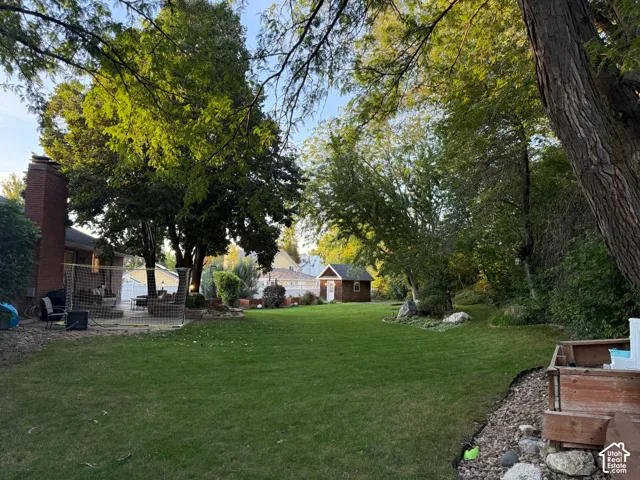 Expansive backyard featuring a manicured lawn, mature trees, a brick chimney, a patio area, and a storage shed