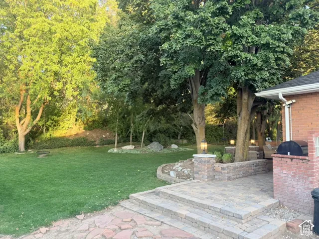 Expansive lawn area with mature trees, brick exterior, multi-level paver patio, stone retaining walls, and built-in outdoor lighting