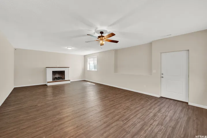 Spacious living area featuring wood-finish flooring, a white brick fireplace with a wood mantle, a window, and a ceiling fan with integrated lighting