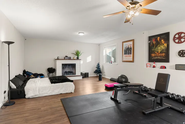 Spacious room with wood-finish flooring, a white-painted brick fireplace, and a ceiling fan with integrated lighting