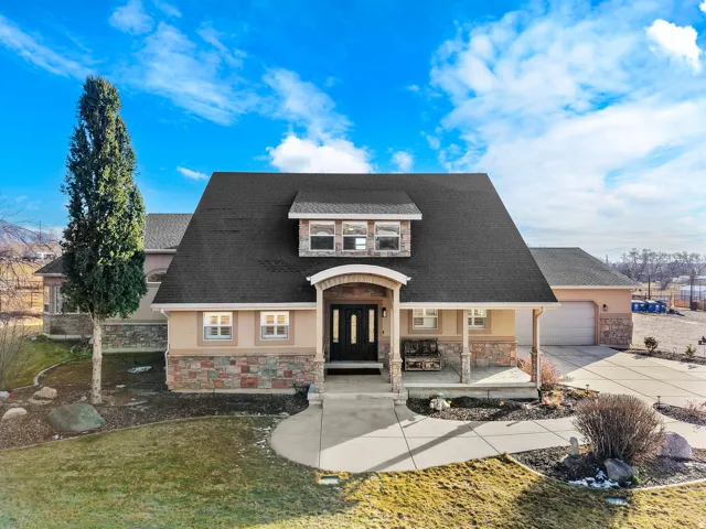 French Colonial-style residence featuring a distinctive mansard roof with dormer windows, cream and stone exterior facade, arched entry portico with black doors, attached two-car garage, manicured lawn with mature landscaping, and mountain backdrop