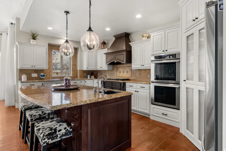 Chef's kitchen featuring white cabinetry with brushed nickel hardware, dual wall-mounted ovens, a six-burner gas cooktop with custom hood, and a large center island with granite countertop and seating for multiple guests