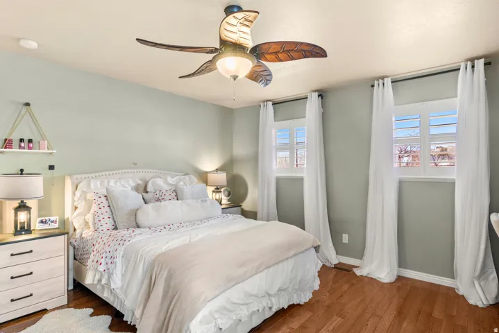 Serene primary bedroom featuring soft sage-green walls, hardwood flooring, and abundant natural light from dual windows with white curtain panels