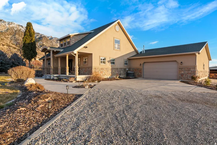 Stucco exterior with stone accents, covered front porch, attached garage, concrete driveway, and gravel parking area