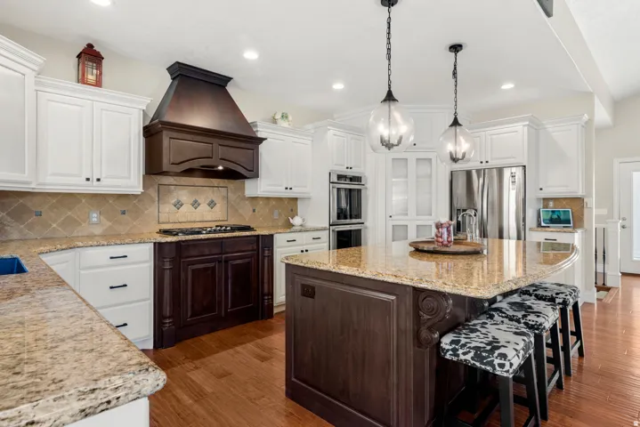 Spacious kitchen featuring an expansive island with granite countertops, stainless steel appliances, white cabinetry, and a dark wood-finish range hood