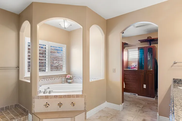 Recessed soaking tub with surrounding tiled accent, arched alcoves, plantation shutters, and a built-in shower bench with mosaic tile