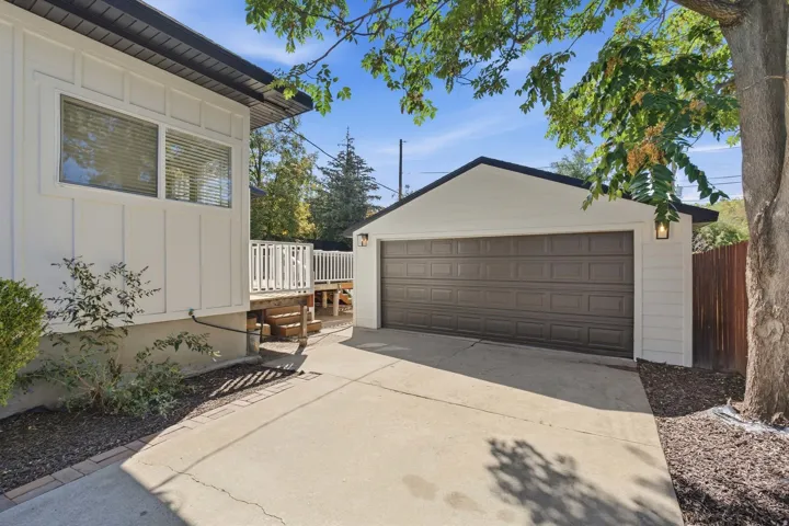 Detached two-car garage with overhead lighting, concrete driveway, white siding, and a wooden deck with railing