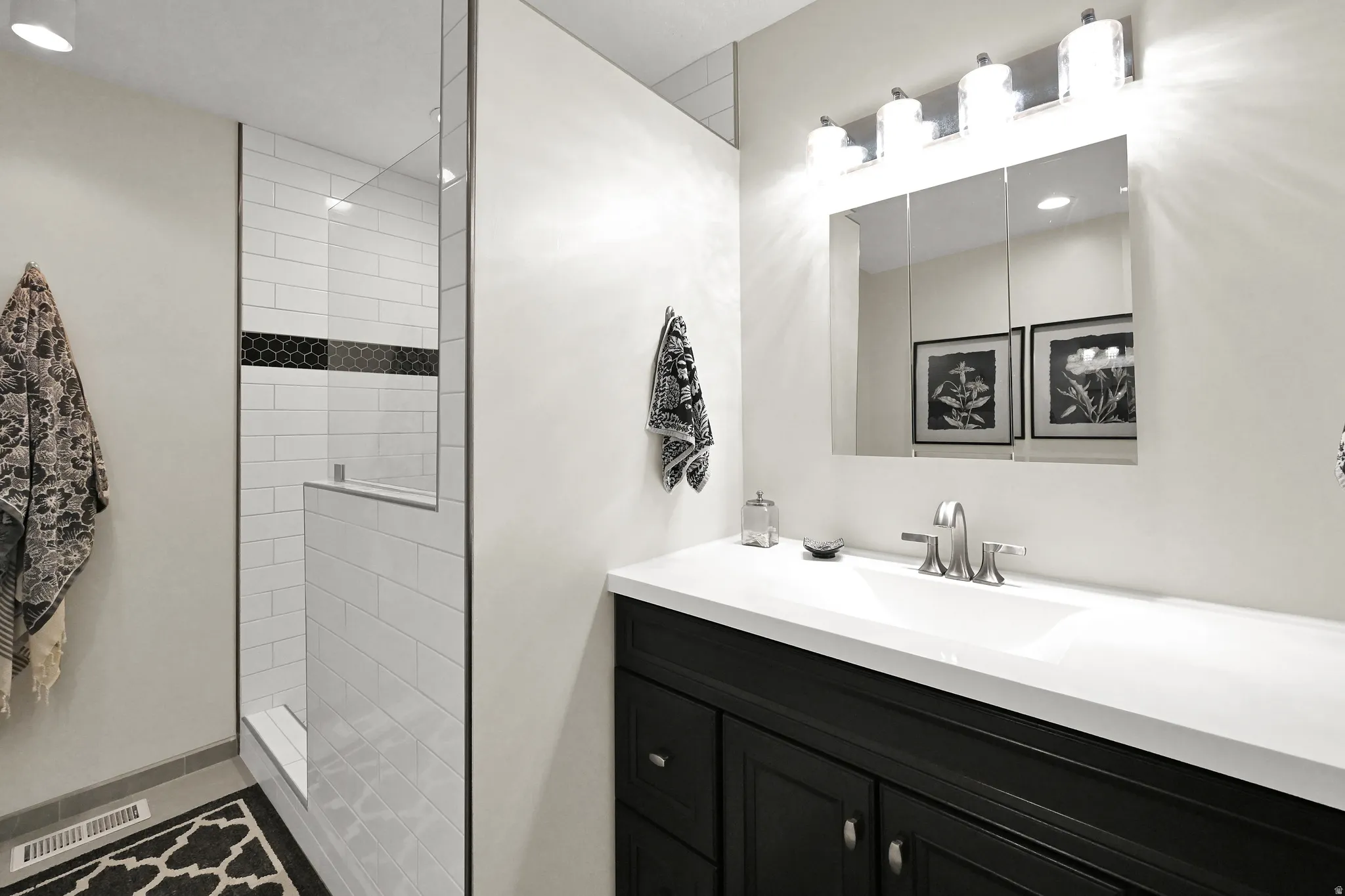 Glass-enclosed shower with subway tile and hexagon accent, complemented by a vanity with a white countertop and dark cabinetry, and a mirrored medicine cabinet