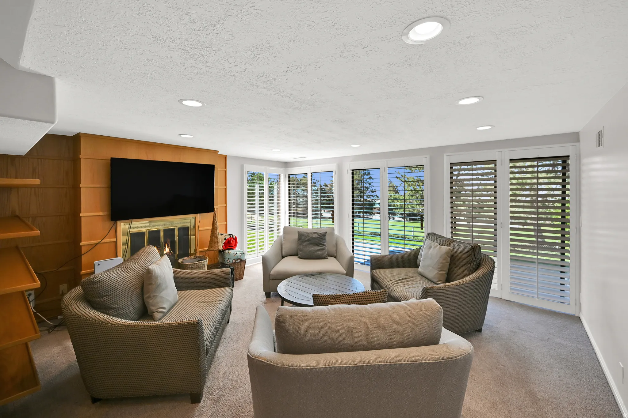 Integrated fireplace with brass-toned detailing, wood-paneled wall, built-in shelving, recessed ceiling lighting, and large windows with plantation shutters