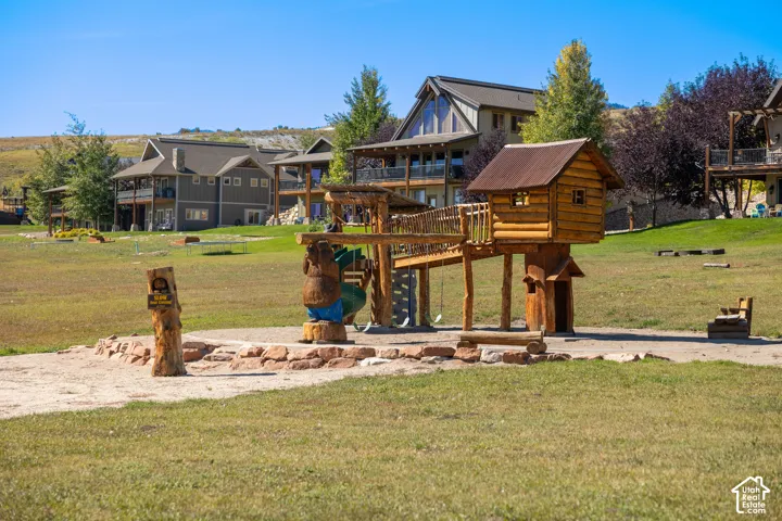 Lush green grounds with a natural wood play structure, a stone-lined fire pit area, and distant residential structures