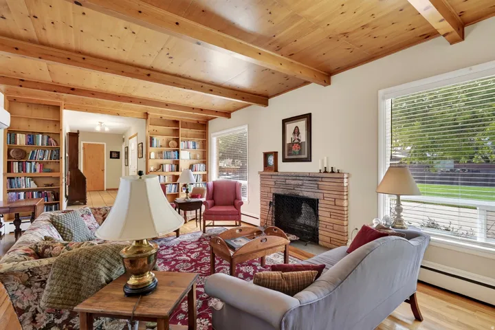 Wood-finish ceiling with exposed beams, stone fireplace, built-in shelving, hardwood flooring, and a large window