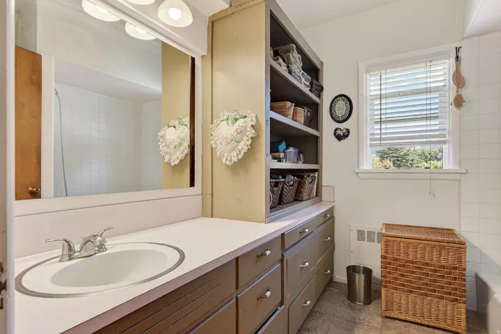 Vanity with an integrated sink and chrome faucet, full-width mirror with overhead lighting, built-in shelving, tiled shower surround, and a window with blinds
