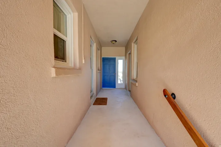 Exterior entry corridor with textured stucco walls, a wooden handrail, and an overhead light fixture leading to a blue entrance door with a sidelight