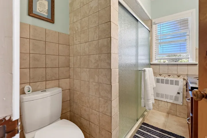 Full-height wall tiling, glass-enclosed shower, white window blinds, and a built-in vanity with dark countertop