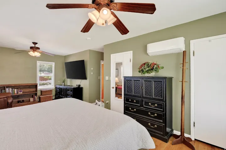 Hardwood flooring, two ceiling fans with integrated lighting, wall-mounted air conditioning unit, and a window with blinds