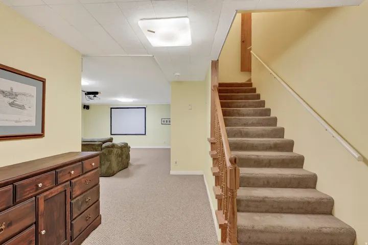 Carpeted staircase with wood banister, recessed ceiling light fixtures, and a projection screen in the background