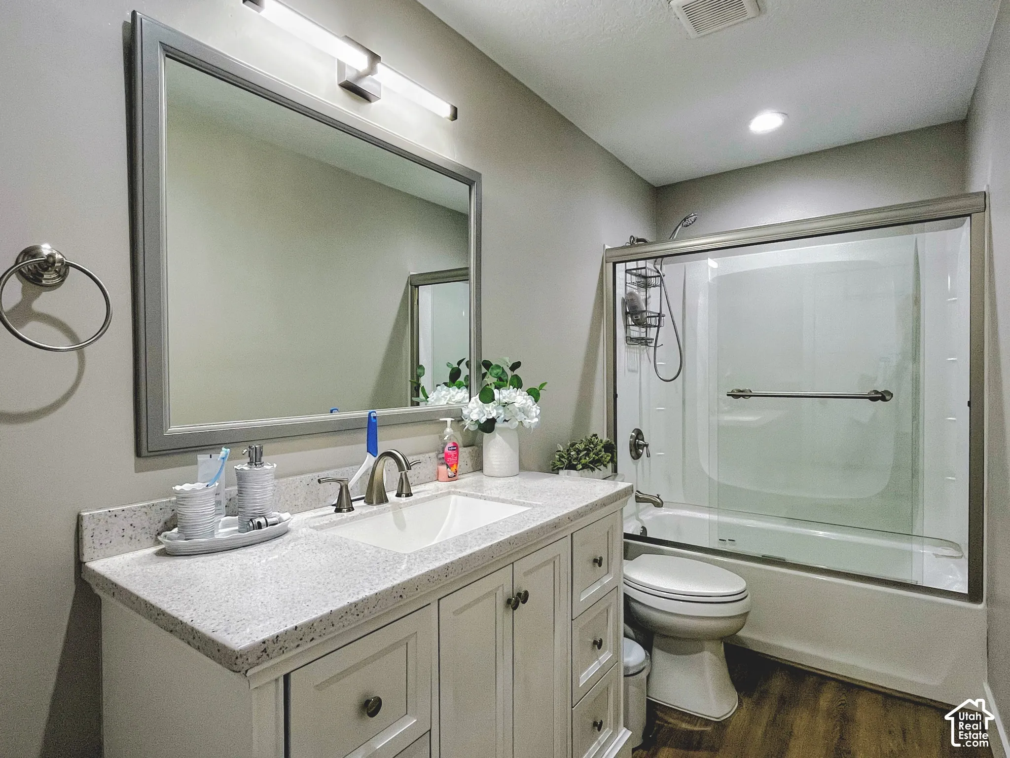 Bathroom with wood-finish flooring, a white vanity with an undermount sink, a stone-finish countertop, and a shower-tub combination with a glass enclosure