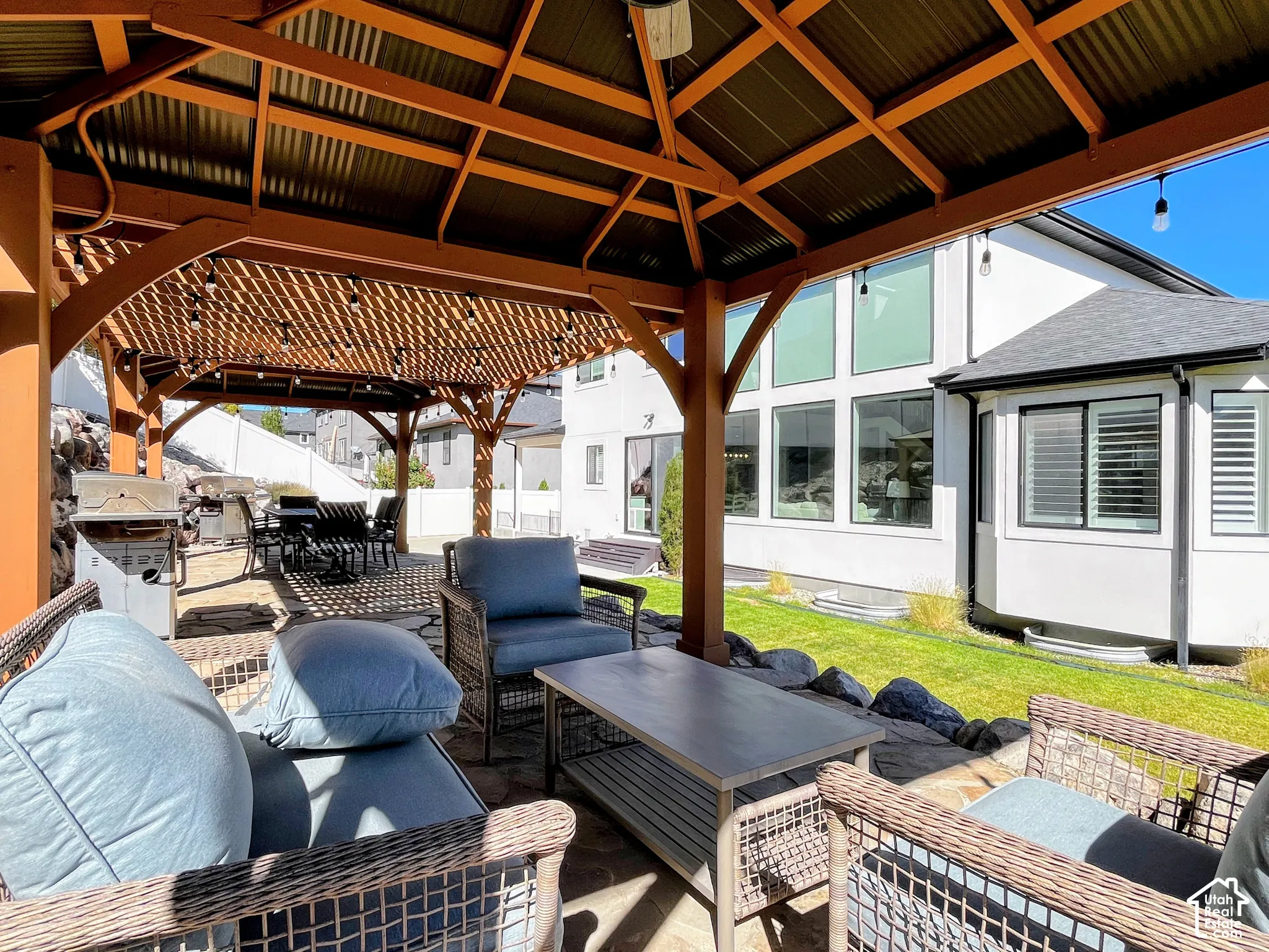 Expansive outdoor living area featuring a covered gazebo with a corrugated roof, integrated lighting, a stone patio, and a built-in outdoor kitchen with a grill