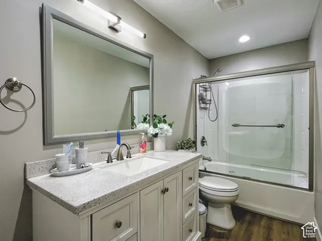 Bathroom with wood-finish flooring, a white vanity with an undermount sink, a stone-finish countertop, and a shower-tub combination with a glass enclosure