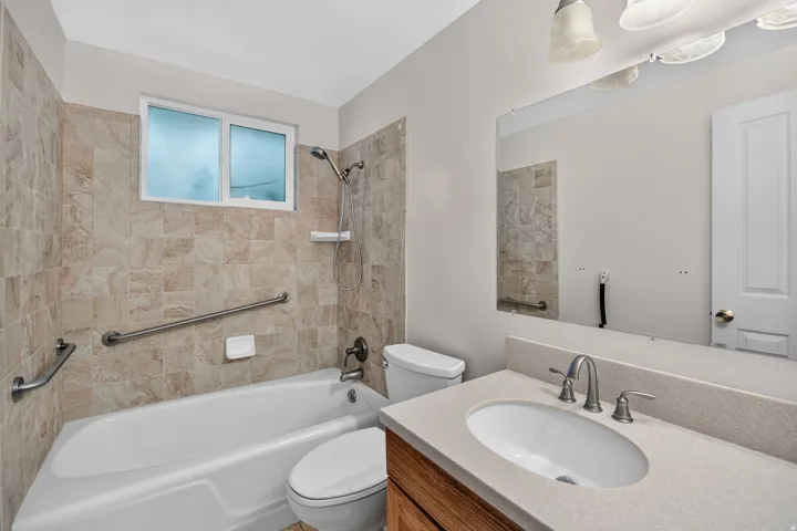 Full tiled shower surround with grab bars, single vanity with light-colored countertop, wall-mounted mirror, and white toilet